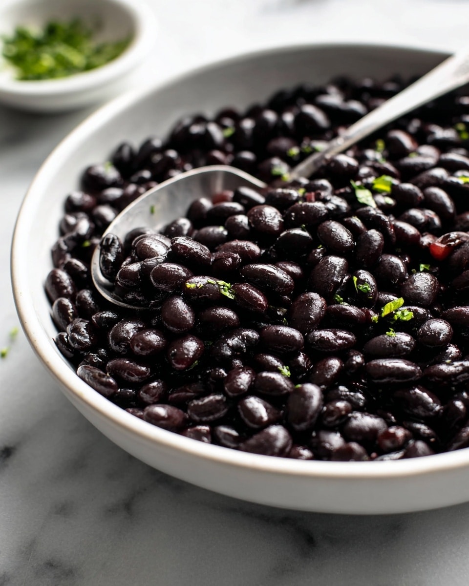 The image shows a close-up of a large white bowl filled with shiny black beans that have a smooth, glossy texture. The beans are densely packed in one layer, with small bits of green herbs sprinkled on top, adding a touch of color. A silver spoon is partially submerged in the beans, with its handle extending out towards the right side of the bowl. The bowl is placed on a white marbled surface, and in the blurred background, there is another small white bowl with green herbs. Photo taken with an iphone --ar 4:5 --v 7