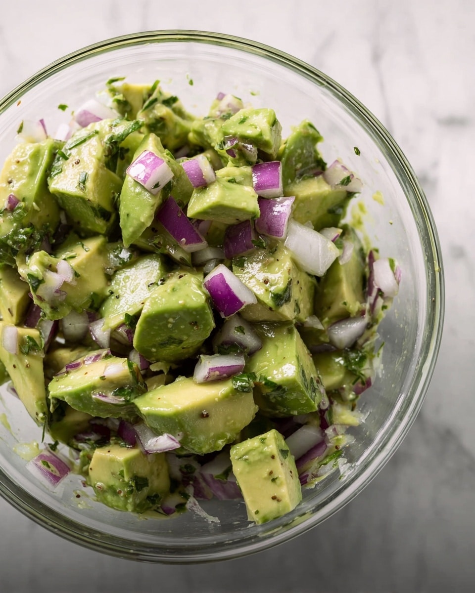 A clear glass bowl filled with chunky pieces of green avocado mixed with small cubes of purple and white onion and some green herbs, all sitting on a white marbled surface. The avocado pieces are soft and uneven, while the onions add a crisp texture with their slightly translucent layers. The mixture looks fresh and lightly coated with a dressing that adds a slight shine. Photo taken with an iphone --ar 4:5 --v 7