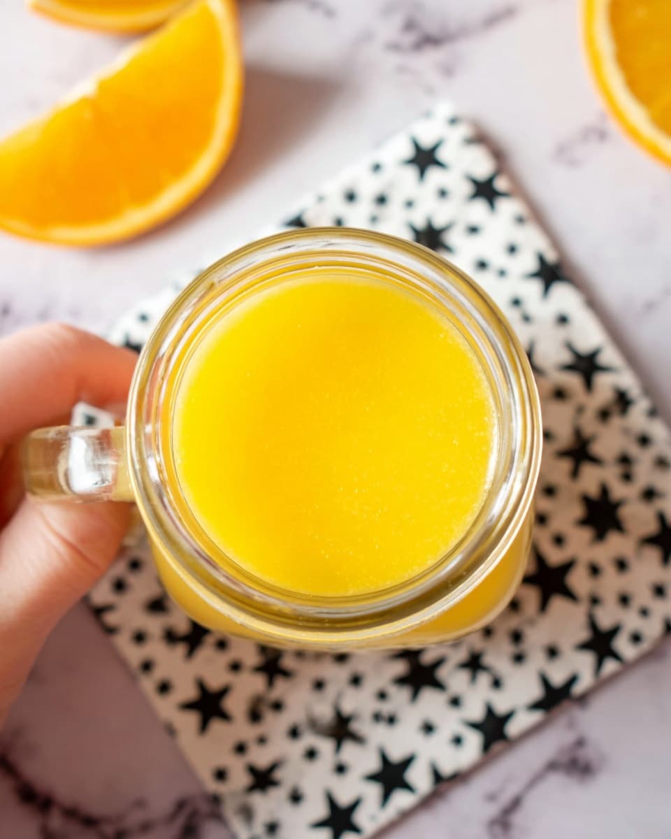 A small clear glass jar filled with a thick, bright yellow-orange liquid, viewed from the top. The jar has a handle on the right, held by a woman's hand. The background shows a white marbled surface with a black and white star-patterned cloth underneath the jar. There are two orange wedges partially visible, positioned on the top left side of the image. The lighting is bright and soft, highlighting the smooth texture of the liquid. photo taken with an iphone --ar 4:5 --v 7