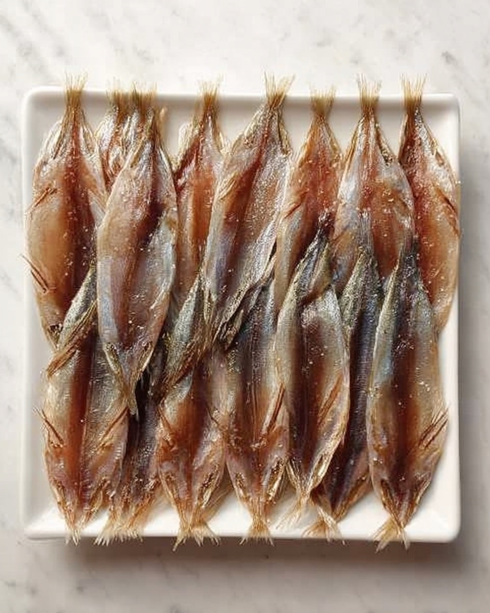 The image shows a long, white rectangular plate on a white marbled surface. On the plate, there are three neat rows of small, flat fish with silvery-brown skin, each fish opened flat like a butterfly with the inside flesh visible. The fish are arranged side by side, with their tails pointing towards the bottom of the plate and their heads towards the top. The skin looks slightly shiny and moist, with a few specks of salt or seasoning visible on the surface. photo taken with an iphone --ar 4:5 --v 7