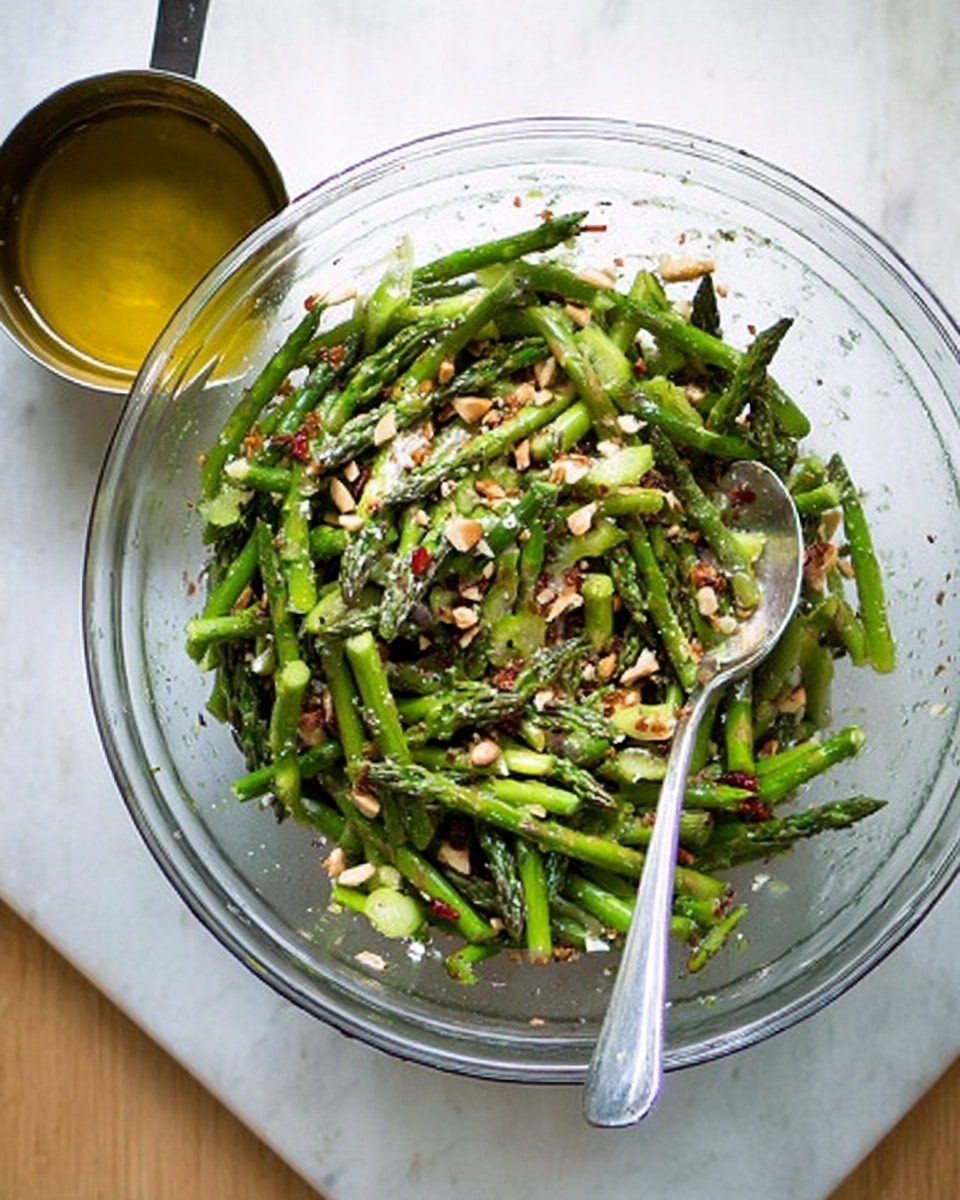 The image shows a clear glass bowl filled with a fresh asparagus salad. The salad has thin green asparagus strips mixed with chopped nuts and red specks that look like chili flakes, all tossed in a light dressing. A metal spoon is resting inside the bowl on the right side. Next to the bowl on the left, there is a small metal measuring cup filled with olive oil. The bowl and cup are placed on a white marbled surface, and the photo is taken from above. photo taken with an iphone --ar 4:5 --v 7