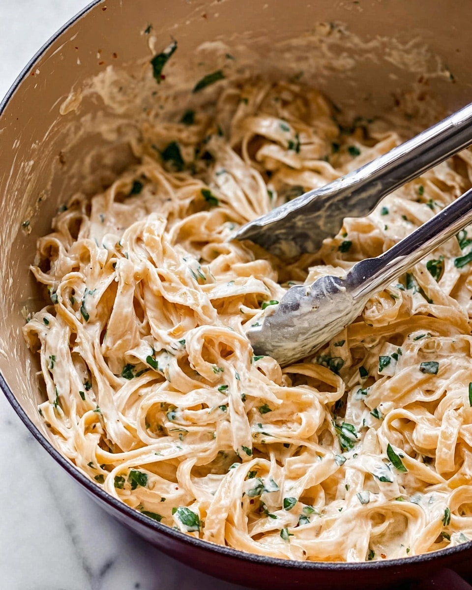 This is a close-up of a pot filled with creamy pasta mixed with small pieces of green herbs. The pasta strands are long and flat, coated evenly in a thick, pale cream sauce that clings to their surface. The green herbs are scattered throughout, adding a speckled contrast to the creamy beige color of the pasta. Two silver tongs are partially visible, resting inside the pot as if stirring the pasta. The inside of the pot is smooth and light colored, while the outside edge has a deep shade that contrasts with the food inside. The scene is set on a white marbled surface. Photo taken with an iphone --ar 4:5 --v 7