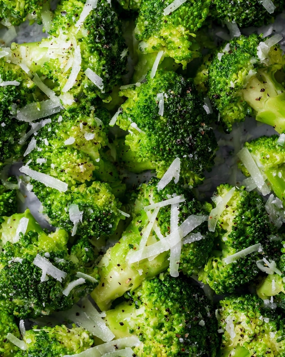 A close-up view of bright green broccoli florets that look fresh and tender, sprinkled with thin, light white shavings of cheese and a light sprinkle of black pepper, all sitting atop a white marbled surface. The green textures of the broccoli are dense, with tiny buds visible on each floret, and the cheese adds a subtle contrast with its fine, translucent flakes spread evenly across the top. photo taken with an iphone --ar 4:5 --v 7