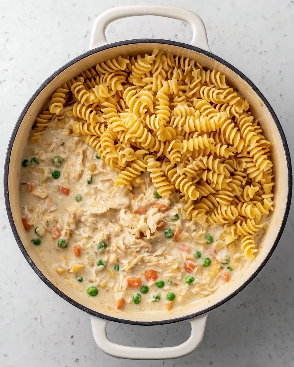 A white pot filled halfway with a creamy sauce containing visible green peas, small orange carrot pieces, and bits of shredded chicken, creating a textured base layer. On top, a generous pile of uncooked yellow spiral pasta covers about half of the sauce, forming a distinct top layer. The pot is placed on a white marbled surface. Photo taken with an iphone --ar 4:5 --v 7