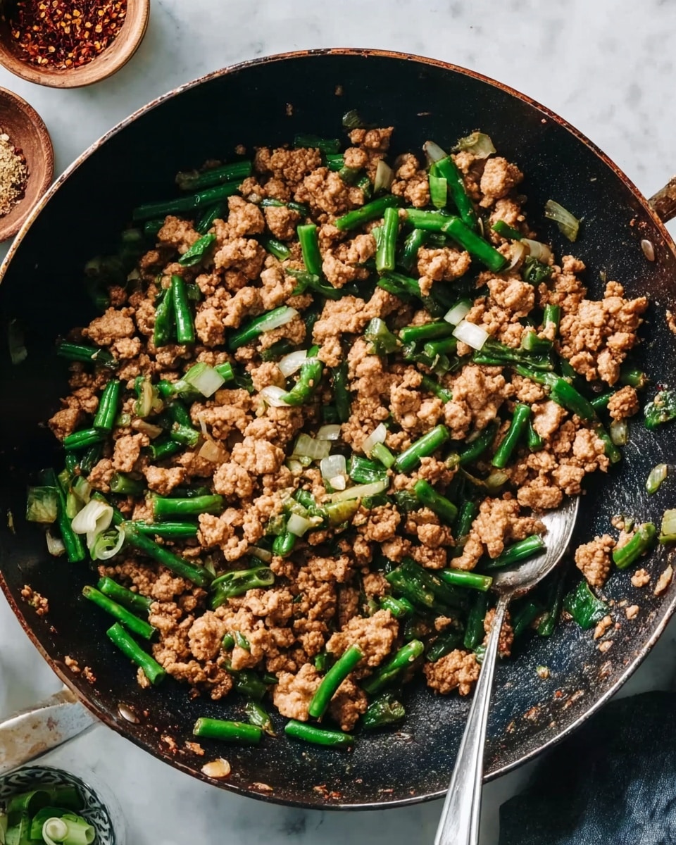A close-up view of a dark pan filled with a stir-fry dish that has two main layers: irregular textured chunks of light brown cooked ground meat and bright green cut vegetable pieces, likely green beans, scattered evenly throughout. Small, diced white onions are mixed in between the meat and vegetables, adding more texture and color contrast. A silver cooking spoon is partially visible inside the pan on the right side, lifting some of the mixture. The background features a white marbled surface with small bowls of spices and a woman's hand holding the pan handle on the right side. Photo taken with an iphone --ar 4:5 --v 7