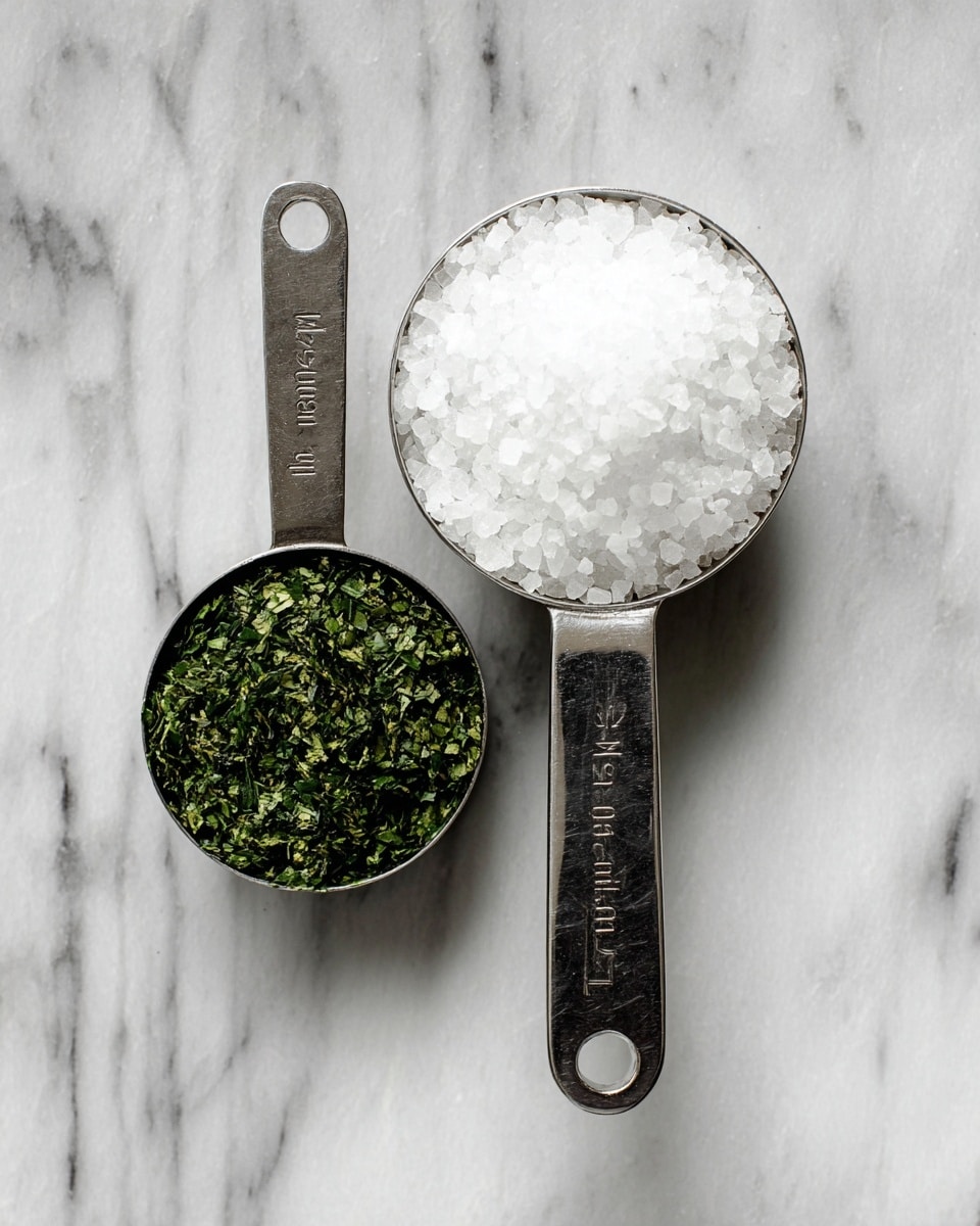 The image shows two metal measuring cups placed side by side on a white marbled surface. The cup on the left contains a half-cup of finely chopped green herbs, with a texture that looks soft and slightly leafy. The cup on the right holds one cup of coarse white salt with a grainy, uneven texture. Both cups have flat handles extending outward, displaying measurement markings that are clearly visible. The overall look is clean and simple, focusing on the contrast between the bright green herbs and the white salt. photo taken with an iphone --ar 4:5 --v 7
