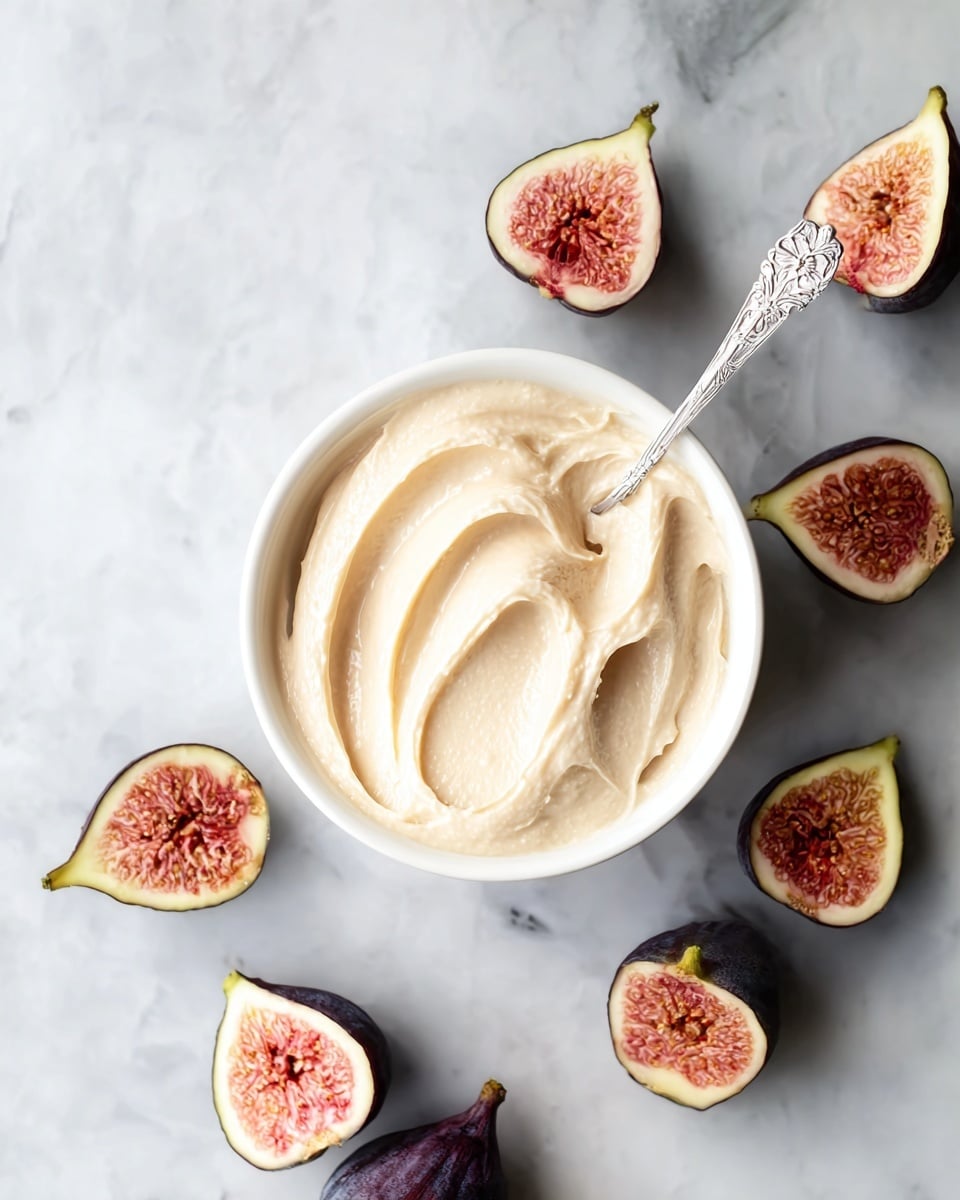 A white bowl filled with smooth, creamy pale beige spread sits in the center on a white marbled surface. A silver spoon with delicate floral details is stuck into the spread, creating soft curved patterns on top. Around the bowl, there are several halves of fresh figs with dark purple skin and bright pink interiors showing detailed seeds, arranged in a loose circular pattern. The figs' inner textures have a mix of soft red, pink, and light yellow tones, contrasting with their dark outer skin. The photo taken with an iphone --ar 4:5 --v 7