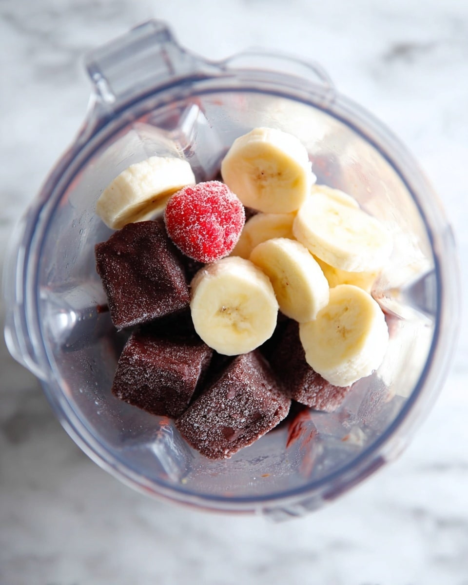 The image shows a clear blender jar filled with ingredients layered inside. At the bottom, there are dark brown frozen cubes with a textured surface, likely açaí or similar fruit puree. On top of the frozen cubes, there are round banana slices, pale yellow with soft edges, evenly spread. Near the bananas, a single small red frozen strawberry with a frosty coating is visible. The blender jar sits on a white marbled surface. photo taken with an iphone --ar 4:5 --v 7