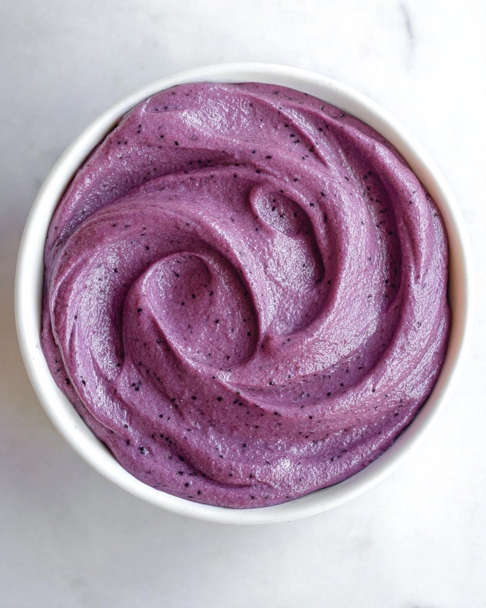 A close-up view of a single layer of thick, smooth, purple spread with a slightly shiny texture, swirled decoratively inside a round white bowl. The spread looks creamy with small specks throughout, giving it a rich, dense appearance. The bowl sits on a white marbled surface that adds a clean and bright background to the vibrant purple color of the spread photo taken with an iphone --ar 4:5 --v 7