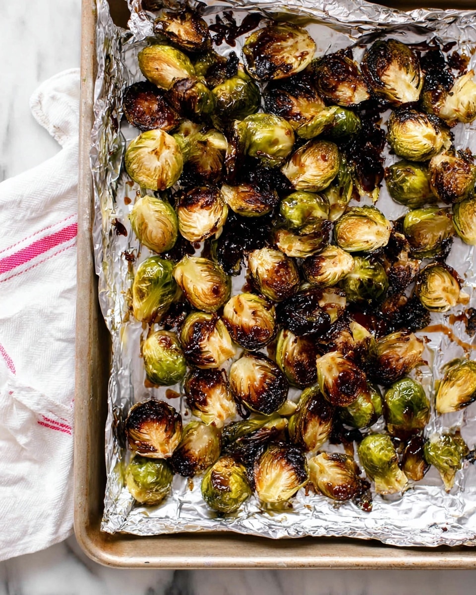 This image shows a baking tray lined with crumpled aluminum foil filled with roasted Brussels sprouts. The Brussels sprouts are halved and arranged loosely in a single layer across the tray. They have a mix of colors from golden brown to dark charred black on the edges and tops, indicating a well-roasted, slightly crispy texture. Some sprouts have a glossy dark brown drizzle, likely a sauce or glaze, scattered unevenly over them. The baking tray is on a white marbled surface, with a folded white cloth featuring red stripes partially visible in the top right corner. photo taken with an iphone --ar 4:5 --v 7
