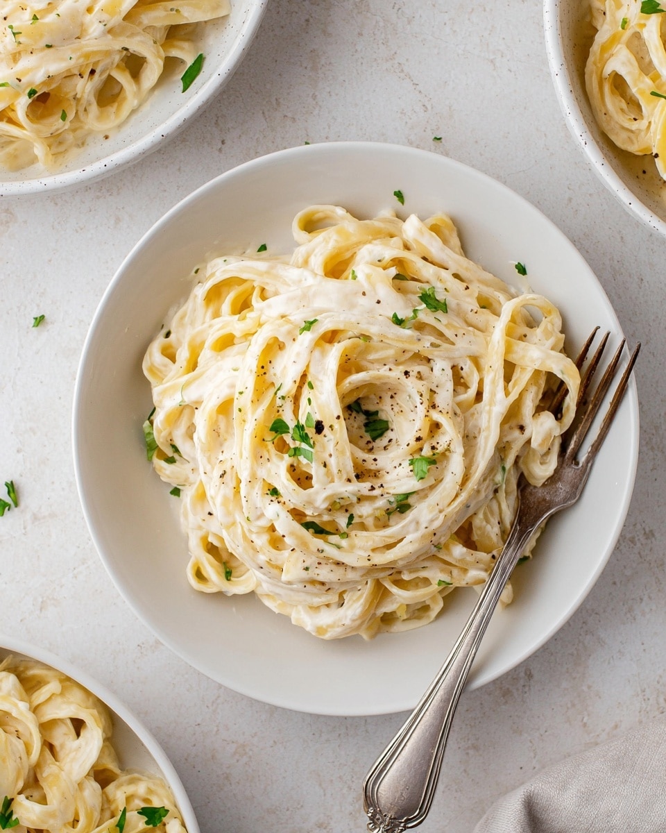 A white plate holds a single portion of creamy fettuccine pasta, with each thick noodle coated evenly in a smooth, pale cream sauce. The pasta is loosely piled in the center, with small green parsley leaves scattered on top and a light sprinkle of black pepper adding contrast. A vintage silver fork rests partially embedded in the pasta on the right side of the plate. Around the main plate, there are partial views of two other white bowls or plates also filled with the same creamy pasta. The whole scene sits on a white marbled surface, giving a clean and fresh look. photo taken with an iphone --ar 4:5 --v 7