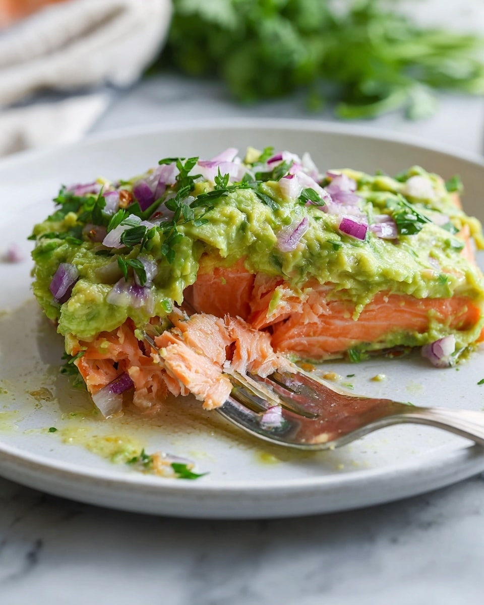 The image shows a close-up of a white plate with a single thick piece of cooked salmon at the center. On top of the salmon is a layer of green mashed avocado mixed with small pieces of red onion and fresh herbs. The salmon's pink-orange flesh is flaky and tender, with some oil glistening on the plate. A metal fork is placed on top of the avocado layer, slightly pressing into the salmon. The plate sits on a white marbled surface with some blurred green herbs visible in the background. photo taken with an iphone --ar 4:5 --v 7