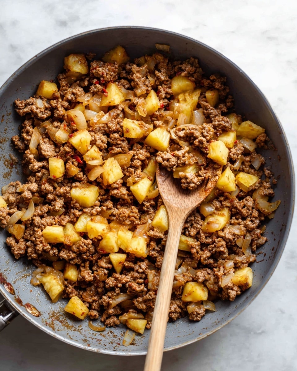 A gray pan filled with cooked ground meat mixed with small chunks of yellow pineapple and pieces of translucent cooked onions, all lightly browned and mixed evenly. A wooden spoon is resting inside the pan, slightly lifting some of the mixture. The surface beneath the pan is a white marbled texture photo taken with an iphone --ar 4:5 --v 7