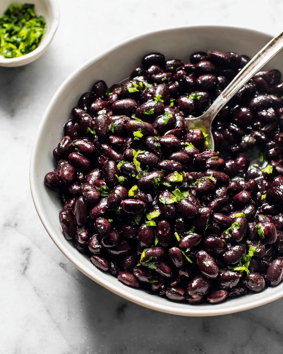 A white bowl filled with shiny, cooked black beans that have a deep dark purple-black color and a smooth, slightly wet texture. Small bright green herb pieces are sprinkled mostly on top of the beans, adding a touch of color. A silver spoon is partially inserted into the beans on the right side of the bowl. The bowl sits on a white marbled surface with a small white bowl of green herbs blurred in the background. photo taken with an iphone --ar 4:5 --v 7