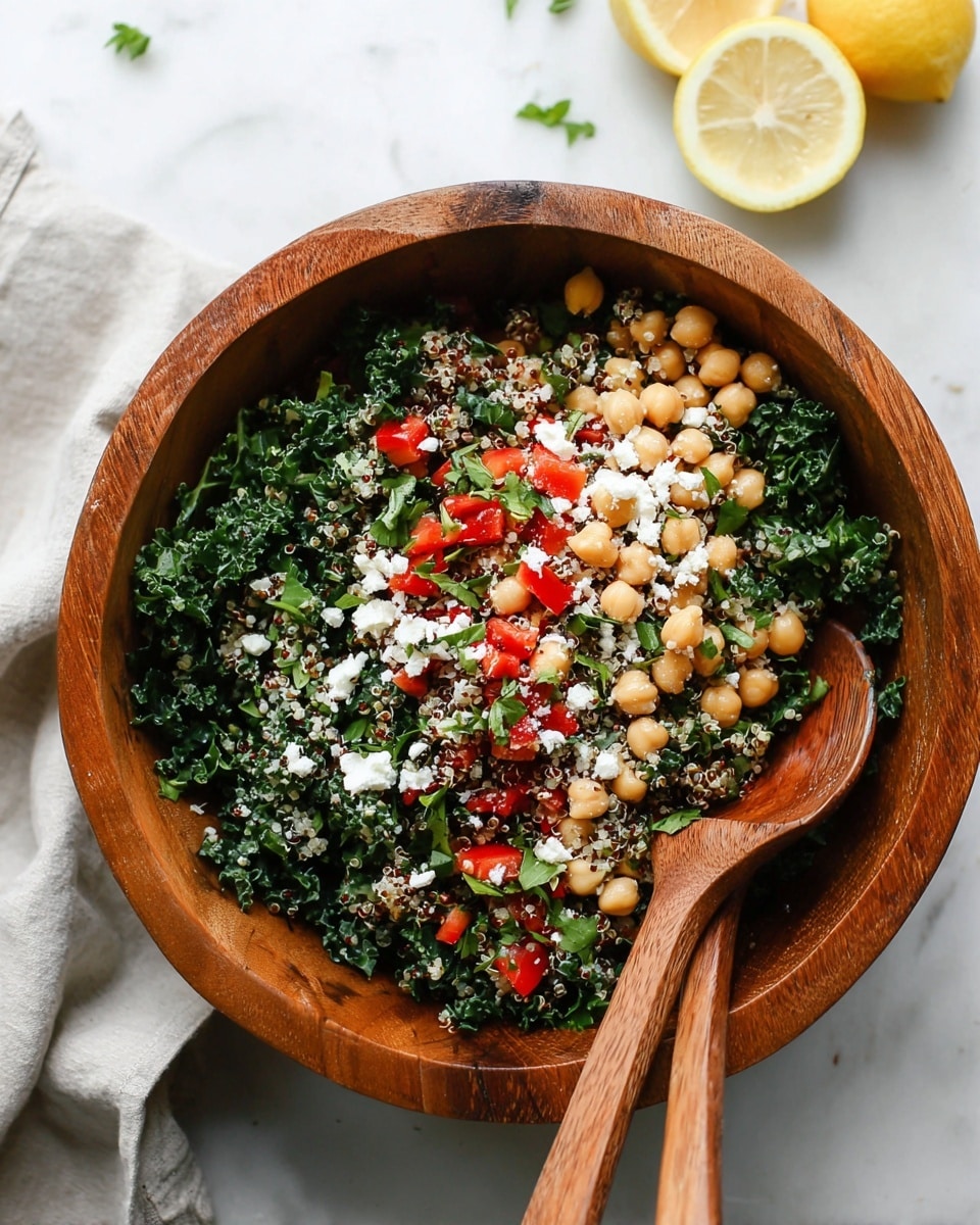 A wooden bowl filled with a fresh salad that has three main layers: a base of dark green curly kale leaves, a middle layer of round beige chickpeas, and a mix of small red diced bell peppers with white crumbled cheese scattered on top. Small bits of quinoa and chopped fresh green herbs are mixed throughout. Two wooden salad spoons rest inside the bowl on the right side. The bowl sits on a white marbled surface with two lemon halves in the top background and a folded light cloth near the bottom left corner. Photo taken with an iphone --ar 4:5 --v 7