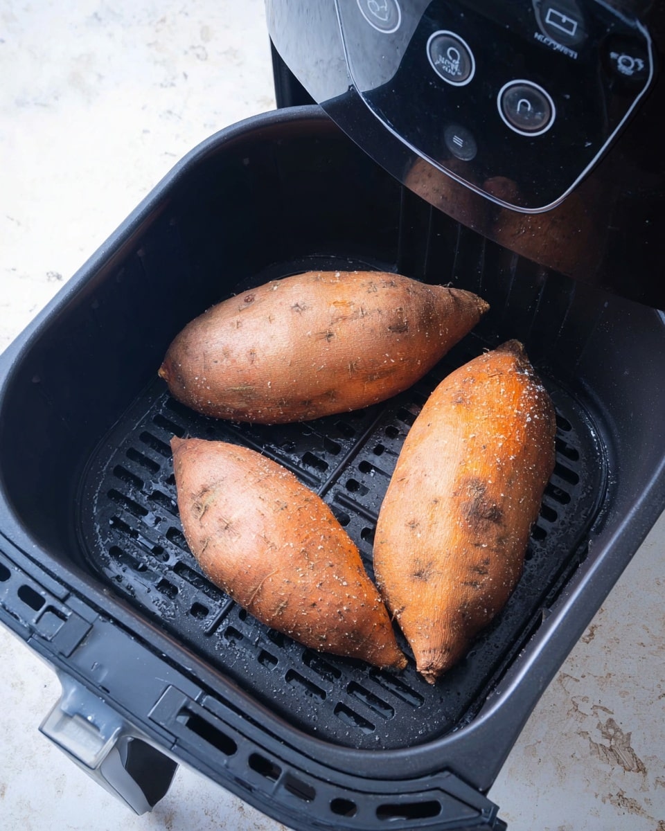Three whole sweet potatoes with rough orange-brown skin are placed inside a black air fryer basket. The basket has a grid pattern for air flow and sits inside the black air fryer machine with a shiny black control panel featuring round touch buttons at the top. The scene is set on a surface with white marbled texture that is only partly visible around the air fryer. The sweet potatoes are arranged in a loose triangle shape with some sparkle from salt or seasoning on the potato skins. photo taken with an iphone --ar 4:5 --v 7