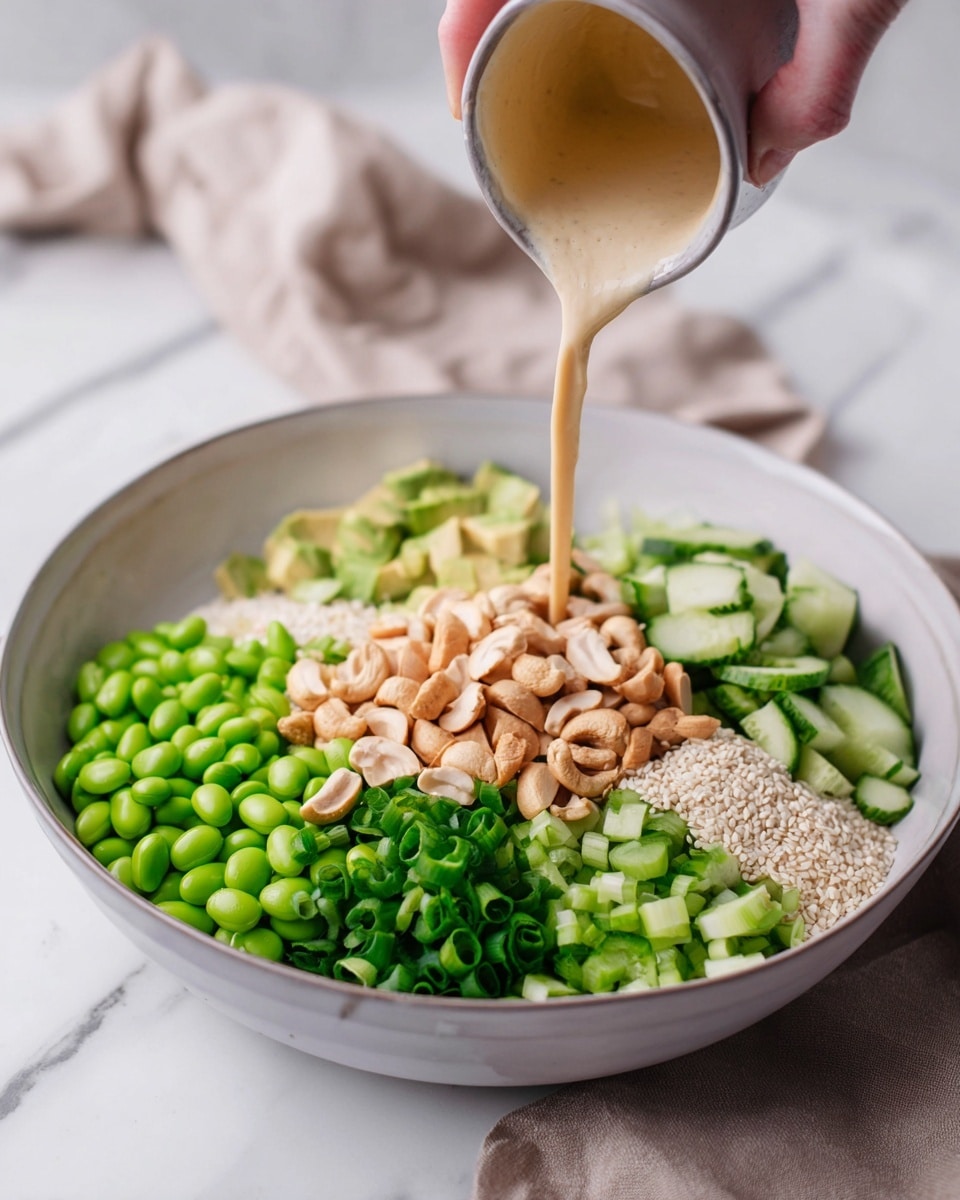 A large white bowl holds a colorful layered salad arranged in neat sections. Starting from the bottom left and moving clockwise, bright green edamame beans form the first layer, followed by chunks of light green avocado, a pile of pale cashew nuts and other nuts, a small mound of white sesame seeds, a heap of chopped dark green spring onions, and a final section of bright green cucumber cubes. A woman's hand is pouring a creamy, light beige dressing over the central pile of nuts. The bowl is set on a white marbled surface with faint gray veins, and a beige cloth is softly blurred in the background. Photo taken with an iphone --ar 4:5 --v 7