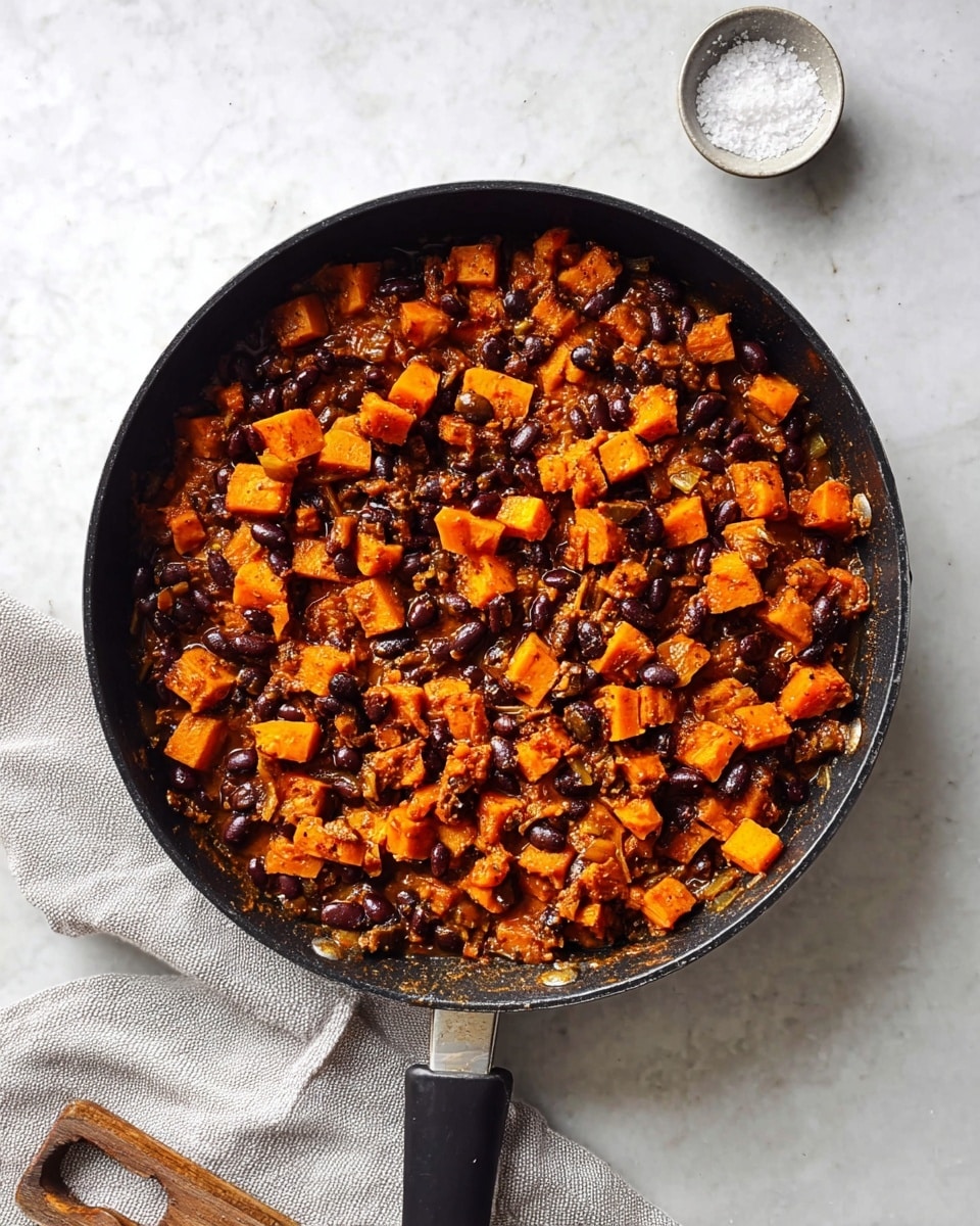 A black skillet filled with a cooked mix of small orange cubes and dark black beans evenly spread inside. The orange cubes are soft with a slightly shiny texture, mixed with the beans soaked in a light brown sauce. The skillet sits on a white marbled surface, with a light gray cloth partially visible on the lower left side and a small white bowl of coarse salt on the lower right. The skillet handle is black and extends toward the bottom center of the image. photo taken with an iphone --ar 4:5 --v 7