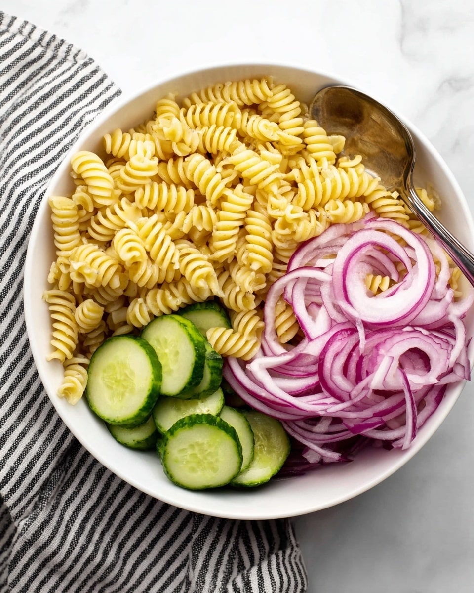 A white bowl sits on a white marbled surface with cooked spiral pasta filling half of it on the right side. On the left side, thin slices of purple-red onion with white inner layers sit at the bottom, while small round green cucumber slices fill the top half. A silver spoon rests next to the bowl on the left, over a black and white striped cloth. The colors are fresh and bright with good natural light photo taken with an iphone --ar 4:5 --v 7
