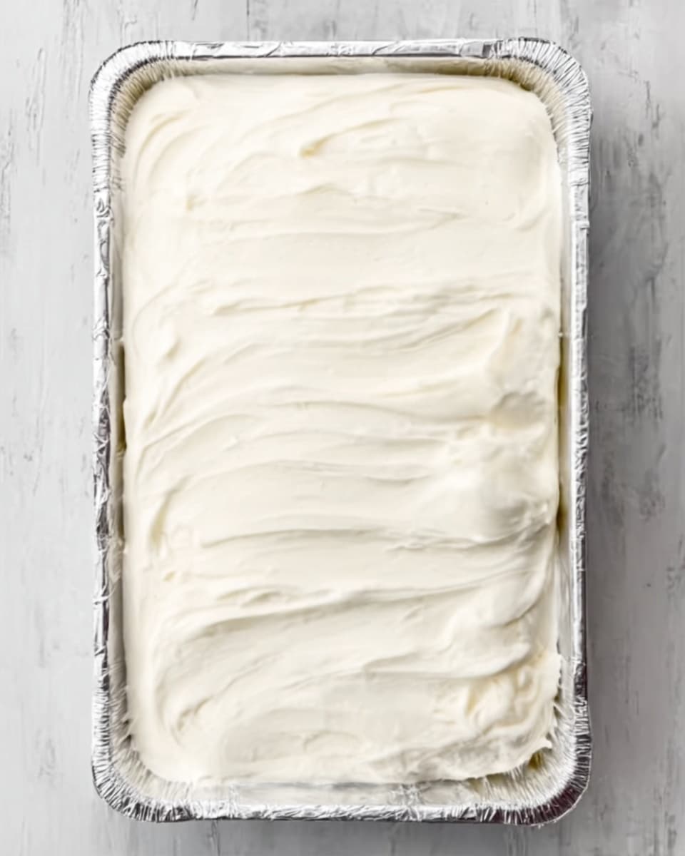 A rectangular foil tray filled with a smooth, thick, creamy white layer evenly spread across the entire surface with visible soft ridges from spreading, placed on a white marbled surface photo taken with an iphone --ar 4:5 --v 7
