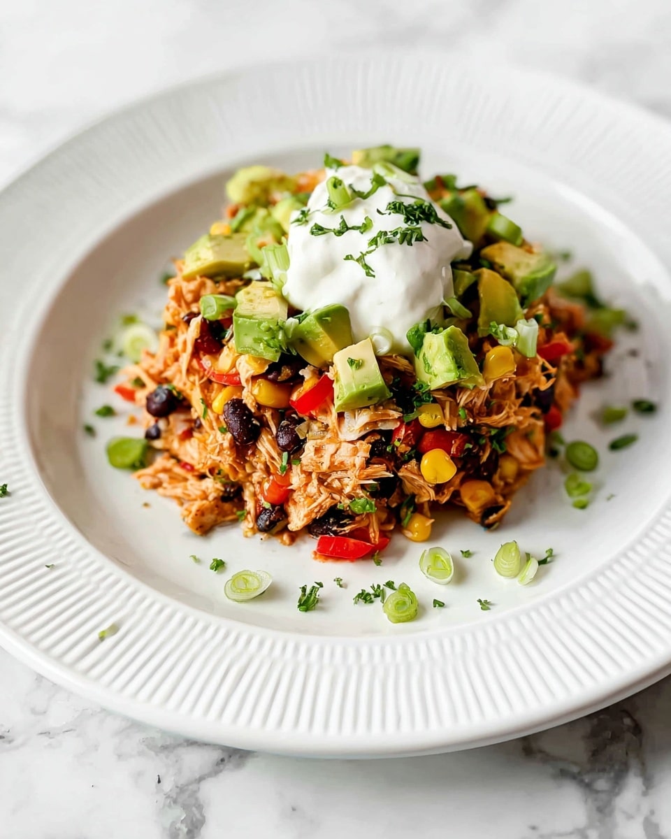 A white ridged plate holds a colorful layered dish with shredded light orange chicken mixed with black beans, yellow corn, and small red pepper pieces, topped with chopped green avocado cubes and sliced green onions. A dollop of white creamy sauce sits in the center of the mound. Small green herb sprinkles add a fresh touch around the plate. The whole scene is set against a white marbled surface. photo taken with an iphone --ar 4:5 --v 7