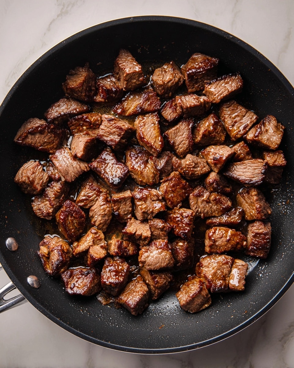 A black frying pan filled with many small pieces of cooked beef, browned and slightly shiny from oil, each piece varying in size and showing a slightly crispy texture on the outside. The pan is placed on a white marbled surface, highlighting the rich brown color and texture of the meat pieces, with some parts darker from cooking. photo taken with an iphone --ar 4:5 --v 7