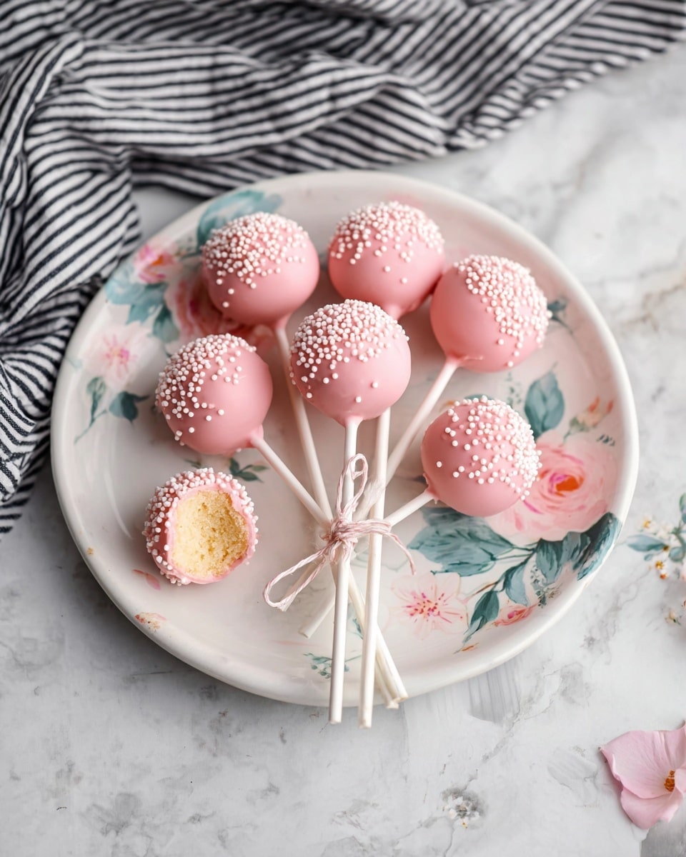 There are eight pink cake pops with a smooth coating and small white round sprinkles on top, arranged in a small bunch tied with a light-colored string on a white plate decorated with pastel pink flowers and green leaves. Two more cake pops lie beside the plate on a white marbled surface, one of them with a bite missing revealing a light yellow soft center. In the background, a black and white striped cloth is draped casually. photo taken with an iphone --ar 4:5 --v 7