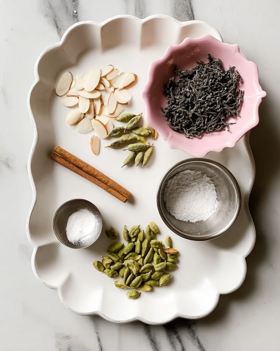 A white tray with a scalloped edge shows six different ingredients arranged neatly: sliced white almonds in the top left section, green cardamom pods near the center, a small brown cinnamon stick laying horizontally below the cardamom, a tiny mound of white powder on a small metal spoon on the left side, a small metal container filled with white granulated sugar in the lower right section, and a cluster of bright green slivered pistachios at the bottom left. To the top right of the tray, a pink bowl holds a pile of dark dried tea leaves. The whole setup sits on a white marbled surface. Photo taken with an iphone --ar 4:5 --v 7