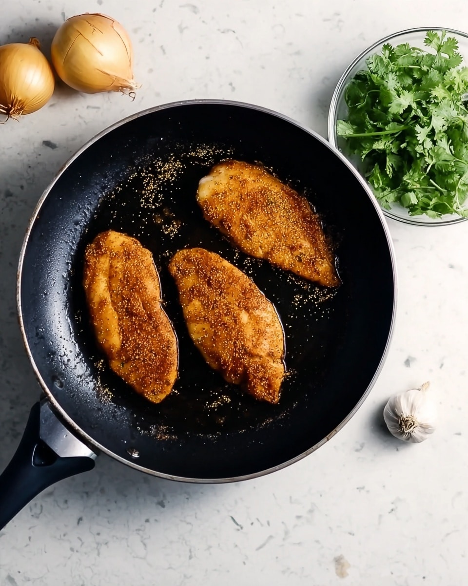 Three pieces of orange-brown seasoned chicken fillets are cooking in a black pan on a stove. The fillets have a slightly crispy texture with some small seasoning dots on them. To the top left of the pan, there are two whole yellow onions and a bunch of fresh green cilantro in a small bowl. To the bottom right of the pan, there is a clove of garlic. The pan and ingredients are placed on a white marbled surface. Photo taken with an iphone --ar 4:5 --v 7