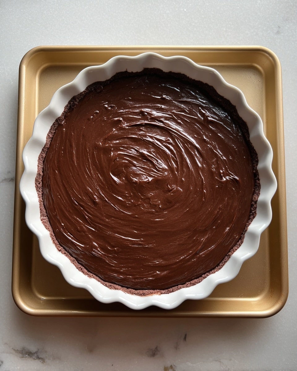 A white round pie dish with scalloped edges holds a single thick layer of smooth, dark chocolate batter spread evenly inside. The batter has a shiny, slightly textured surface with swirls made from spreading, showing some light reflections. The pie dish rests on a flat, light gold baking tray, and the whole scene is set on a white marbled textured surface. photo taken with an iphone --ar 4:5 --v 7