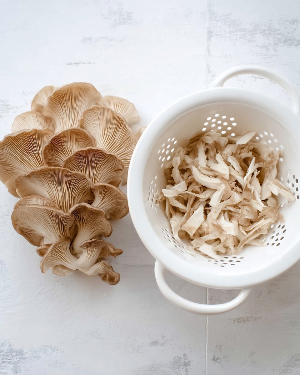 The image shows a cluster of light brown oyster mushrooms with soft, fan-shaped caps and delicate gills on the left side, sitting on a white marbled surface. On the right side, there is a white colander filled with thinly shredded oyster mushroom pieces, showing a mix of cream and beige colors and a soft texture. The colander has large holes and a handle, resting flat on the white marbled surface. Photo taken with an iphone --ar 4:5 --v 7