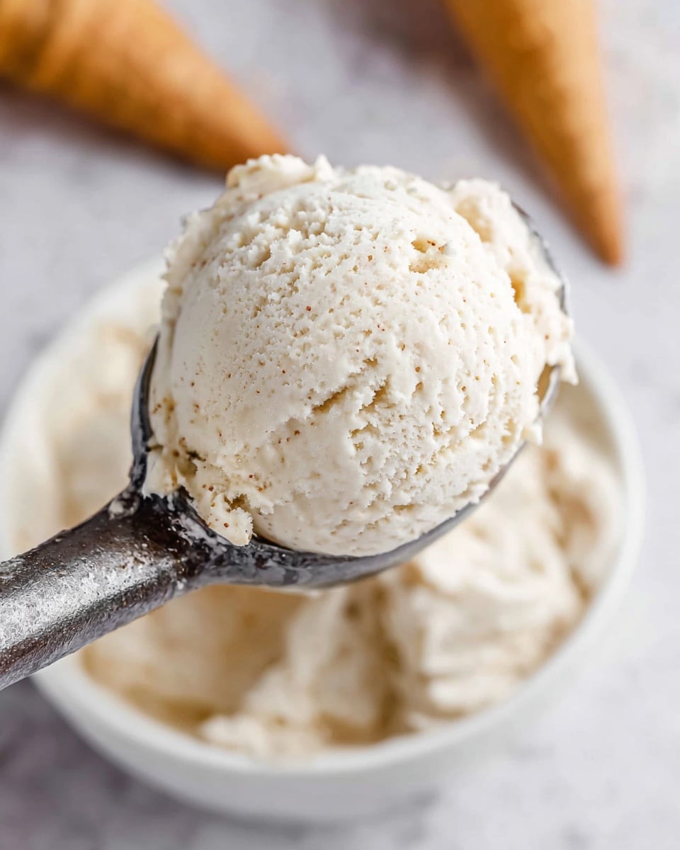 A close-up view of one large scoop of creamy vanilla ice cream with small brown vanilla specks, held on a shiny metal ice cream scooper above a white bowl that also holds more ice cream. The scoop is round and textured with soft ridges showing the smooth, rich ice cream. In the blurred background, a golden-brown ice cream cone is partially visible on a white marbled surface photo taken with an iphone --ar 4:5 --v 7