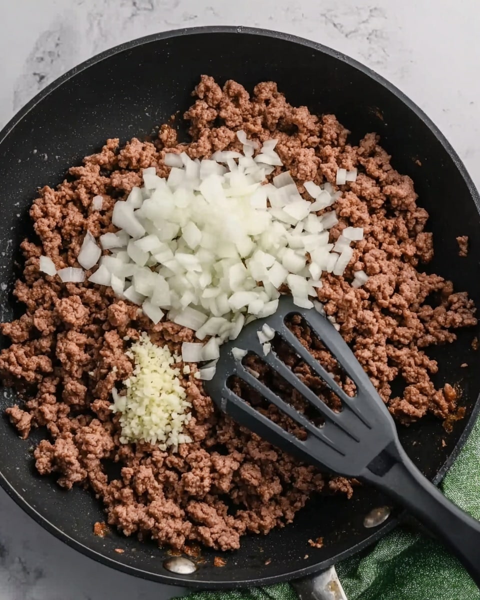 A black skillet filled with cooked ground beef spread evenly. On top of the beef, there is a pile of finely chopped white onions and a smaller pile of minced garlic placed next to each other near the center. A black slotted spatula rests inside the skillet, partly under the beef, on the right side. The skillet is set on a white marbled surface with a green cloth partially visible on the right side. Photo taken with an iphone --ar 4:5 --v 7