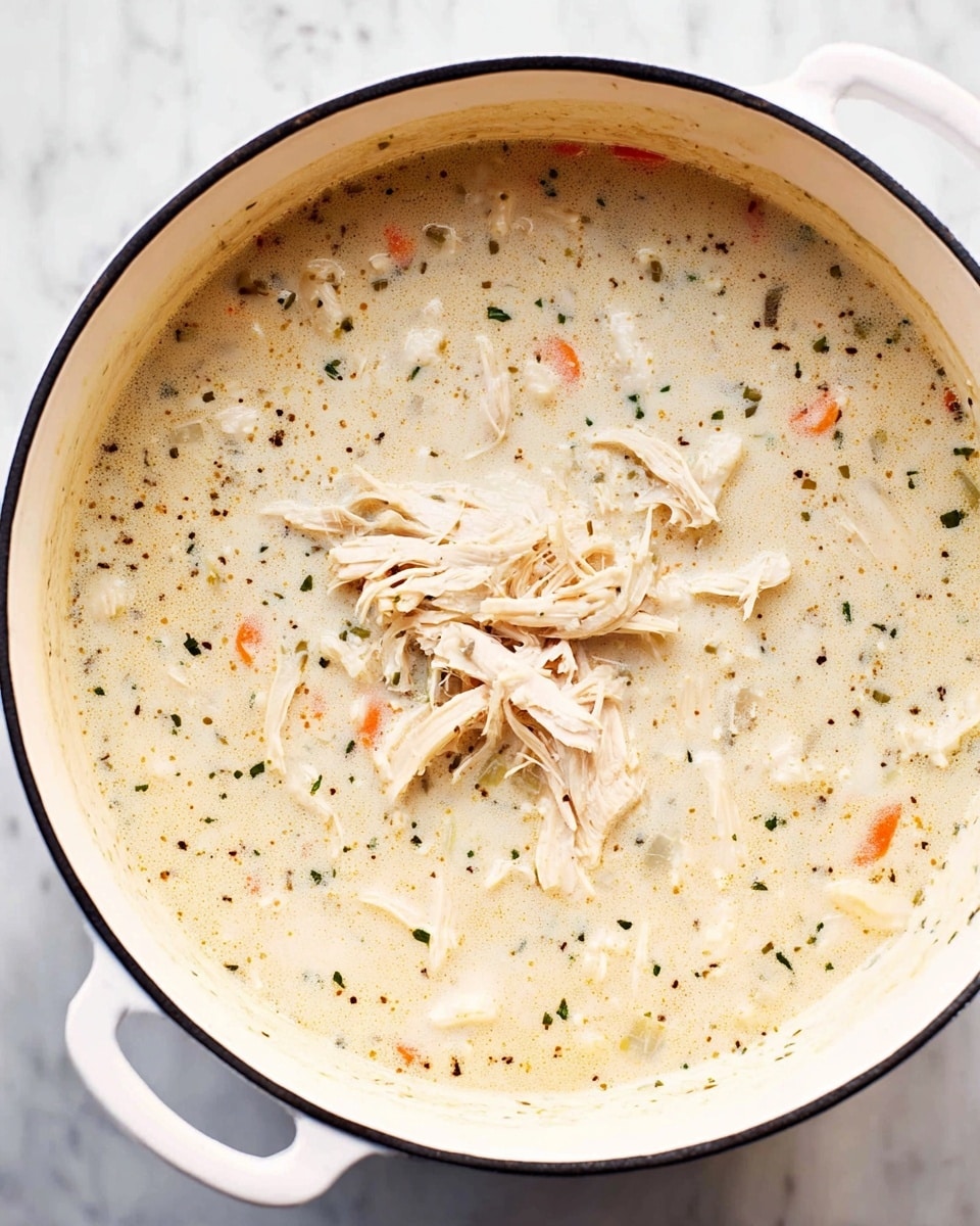 A white pot filled with creamy soup that has a light beige color with small black and green herb specks throughout. Floating on top are several shredded pieces of light beige chicken. Thin strips of orange carrot peek through the soup near the edges. The texture looks thick and smooth with visible bits of vegetables and spices underneath. The pot is placed on a white marbled surface. photo taken with an iphone --ar 4:5 --v 7