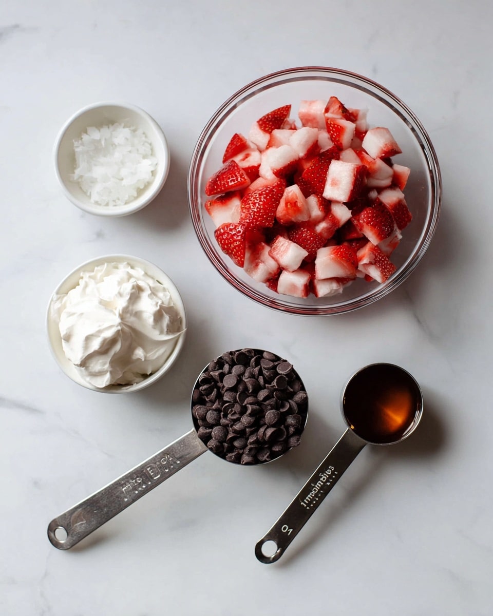 The image shows five different white kitchen containers and measuring spoons set on a white marbled surface. In the center is a clear glass bowl filled with chopped red and white strawberries, showing a mix of bright red and pale pink pieces. To the bottom right, a metal measuring cup overflows with small dark chocolate chips, nearly filling the container. To the left, a metal measuring cup holds smooth white whipped cream with soft peaks. Above the whipped cream, a white bowl contains a small amount of white solid, likely coconut oil, with a soft texture. Two metal measuring spoons lie on the surface; one holds a dark amber liquid and marked