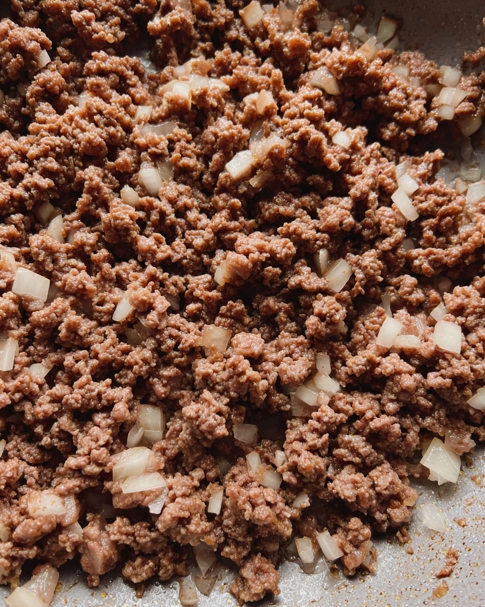 This image shows cooked ground beef mixed with small pieces of cooked white onion in a pan. The ground beef is brown with a crumbly texture, scattered evenly across the pan with bits of translucent onion. The pan's inner surface is visible and has a metallic shine. The background is a white marbled texture. photo taken with an iphone --ar 4:5 --v 7