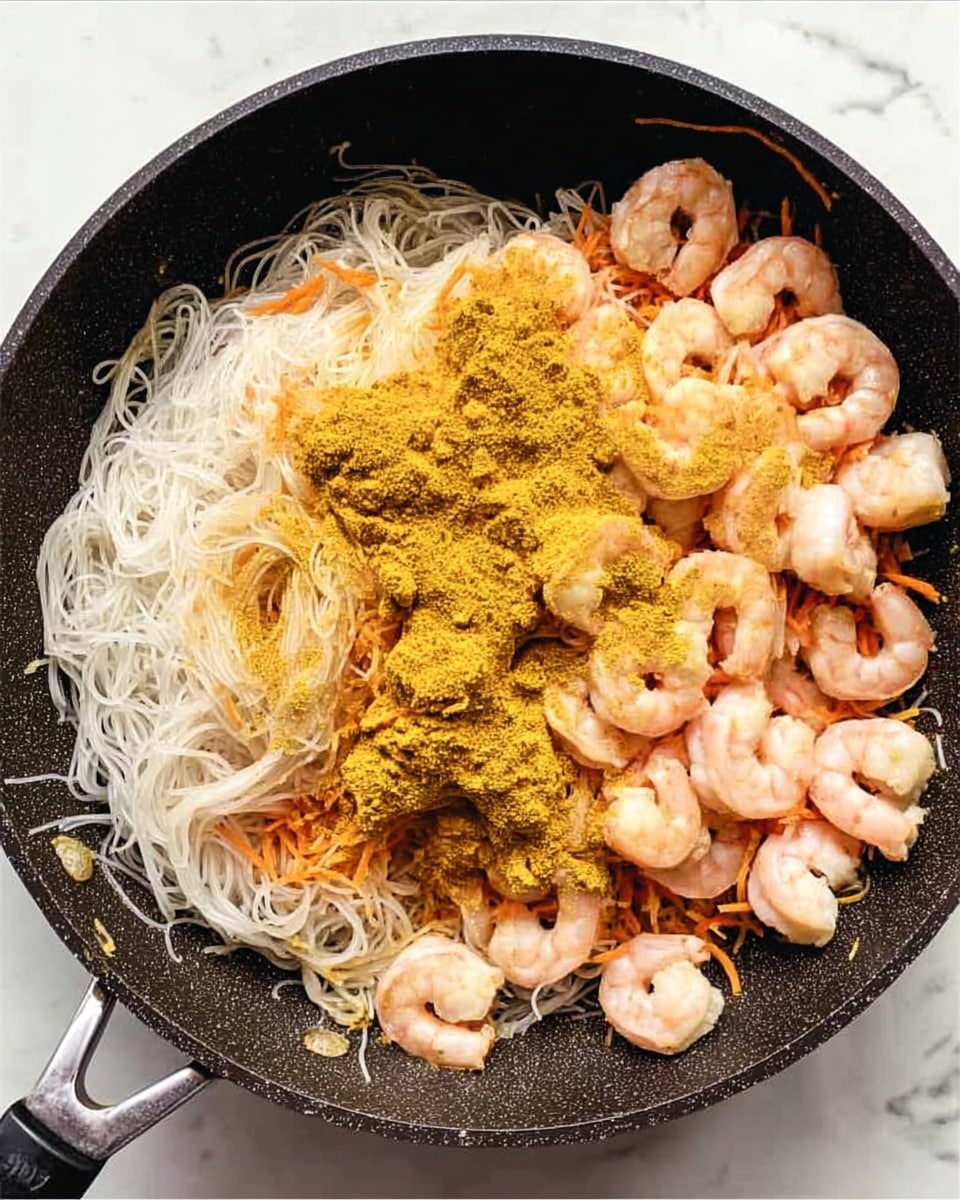 A black pan filled with three main layers: the bottom layer has thin, white rice noodles arranged loosely, the middle layer contains thin orange carrot strands mixed lightly in, and the top layer shows a spread of light pink cooked shrimp scattered unevenly, all covered with a dusting of bright yellow curry powder mostly concentrated in the center. The pan is placed on a white marbled surface. photo taken with an iphone --ar 4:5 --v 7