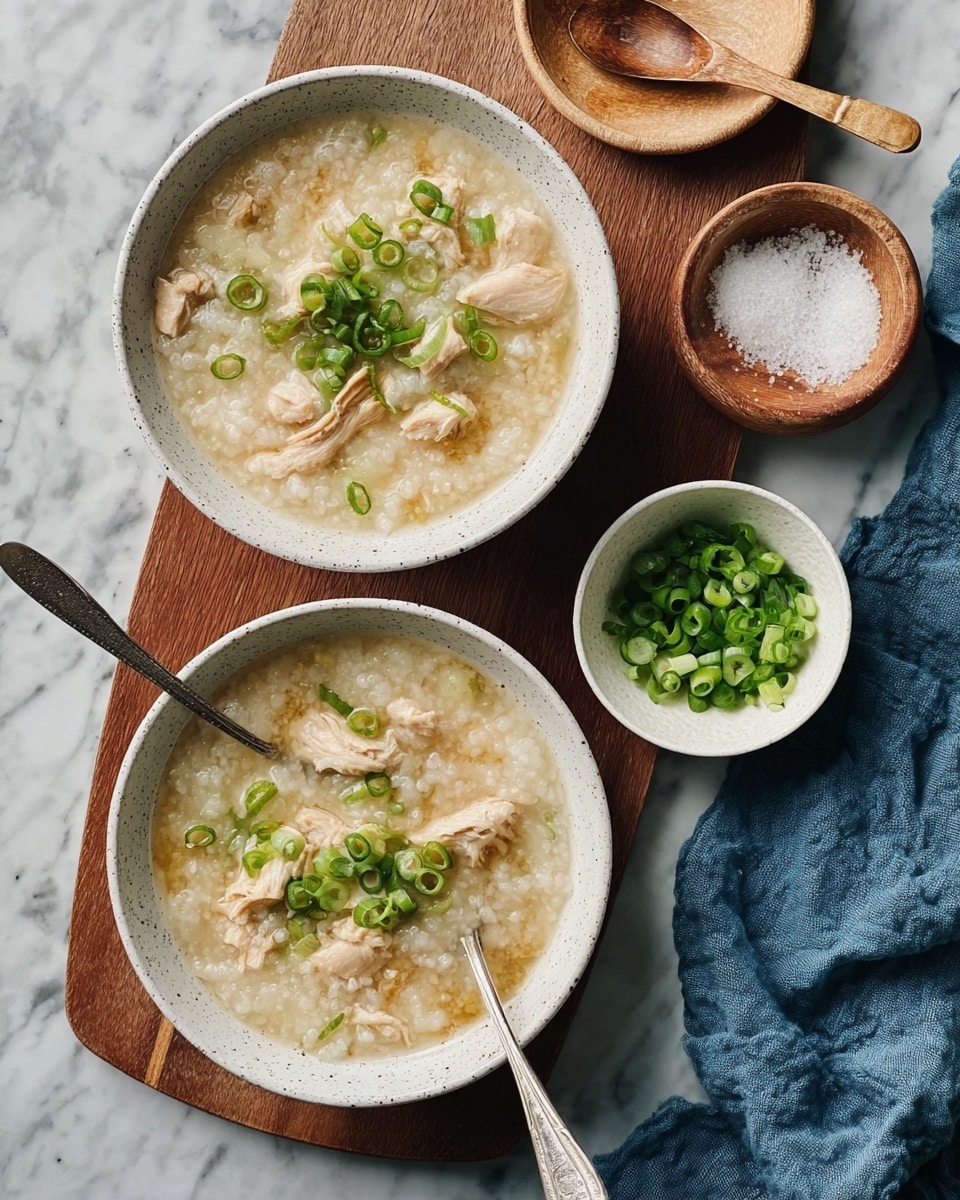 Two bowls of thick rice porridge with soft pieces of light beige chicken mixed in, garnished with small rounds of bright green scallions on top. Each bowl has a silver spoon resting inside. The bowls are white with a speckled texture and sit on a wooden board and a white marbled surface. Next to them, there is a small white bowl filled with chopped green scallions and a small wooden bowl with coarse white salt and a small wooden spoon. A blue cloth napkin is casually placed near the bowls. Photo taken with an iphone --ar 4:5 --v 7