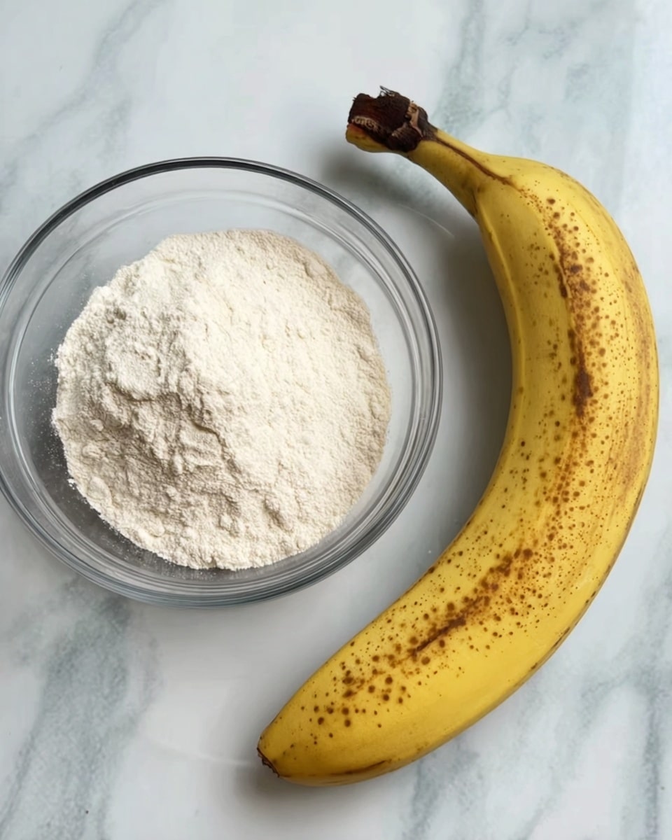 A clear glass bowl filled with white flour sits on a white marbled surface next to a large ripe banana that is yellow with many brown spots, showing its softness and sweetness. The bowl's round shape clearly holds the fine, powdery flour, and the banana curves gently with a darkened stem at the top. photo taken with an iphone --ar 4:5 --v 7