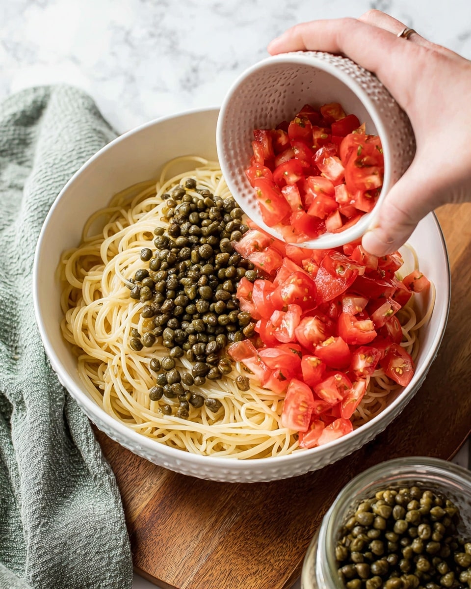 A white bowl on a white marbled surface holds a serving of cooked spaghetti with two visible layers on top: on the left side, a layer of small dark green capers, and on the right side, a layer of bright red diced tomatoes. Above the bowl, a woman's hand is pouring more diced tomatoes from a small textured white bowl into the pasta. In the front right corner, there's a glass jar filled with capers, and to the left, a gray-green cloth napkin is partially visible. The scene is lit naturally, highlighting the fresh colors and textures of the food photo taken with an iphone --ar 4:5 --v 7