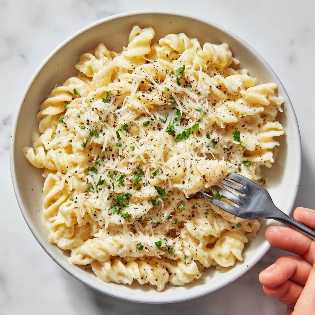 A close-up of creamy pasta served in a white bowl on a white marbled surface, showing two main layers: the pasta which is a pale yellow color with a twisted shape, and the thick creamy white cheese sauce coating it evenly. The dish is topped with fine shreds of cheese, small green parsley bits, and a light sprinkle of black pepper, adding texture and color contrast. The edges of the bowl are slightly visible, framing the rich pasta inside. photo taken with an iphone --ar 4:5 --v 7