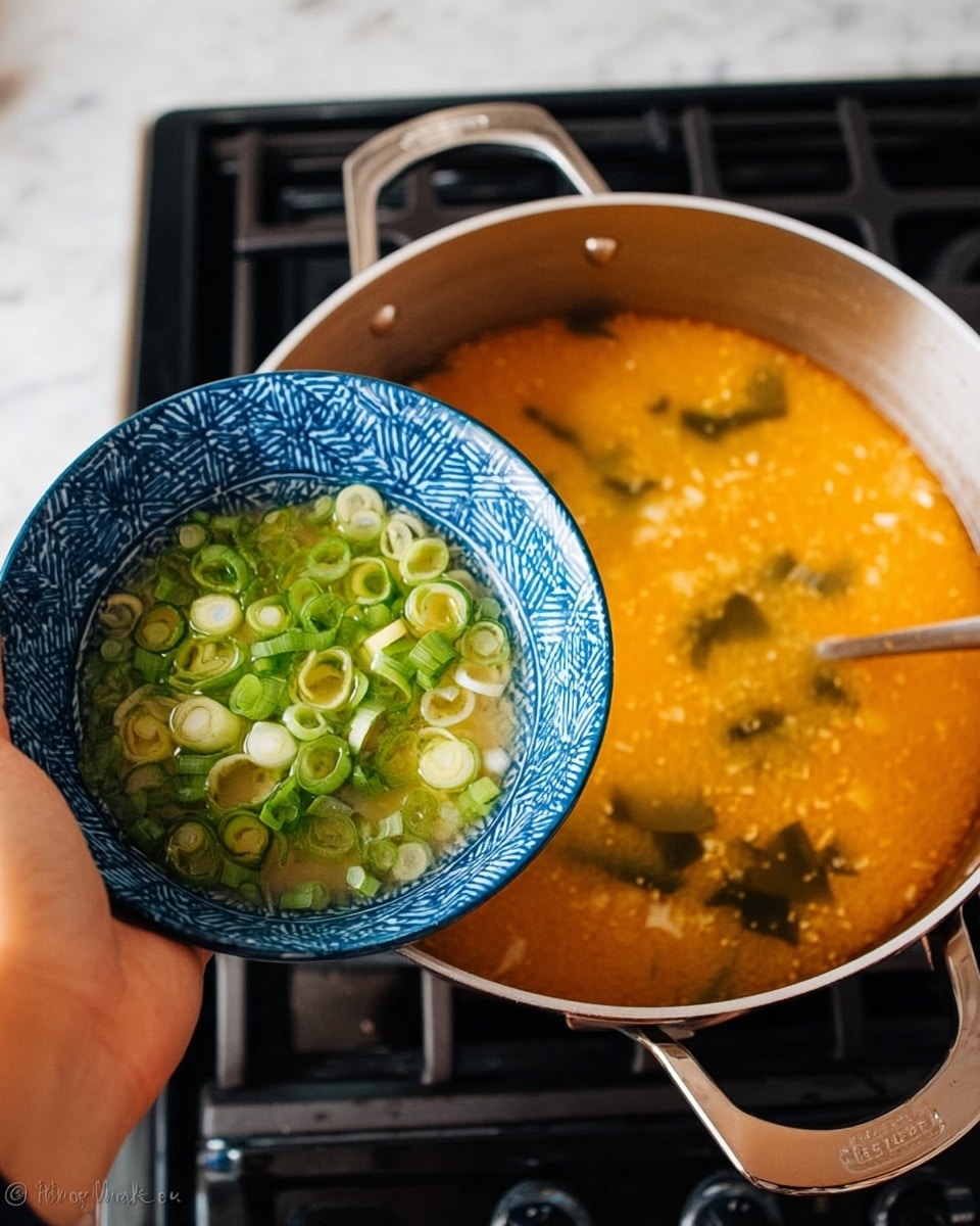 A woman's hand holds a blue and white bowl with sliced green onions over a silver pot of orange miso soup on a stove, with visible seaweed and tofu pieces inside the soup. The background surface is white marbled. photo taken with an iphone --ar 4:5 --v 7