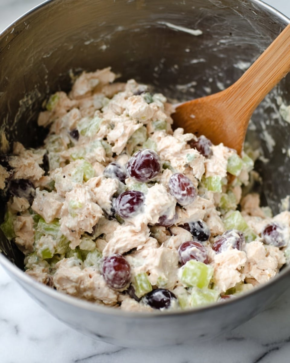 A close-up view of a large metal mixing bowl filled with a creamy chicken salad mixture. The mixture has three main layers: the white creamy base, small chunks of white chicken meat, pieces of light green celery, and dark purple grapes cut in halves. A light brown wooden spoon is partially inside the bowl, mixing the salad. The background surface is white marble. photo taken with an iphone --ar 4:5 --v 7