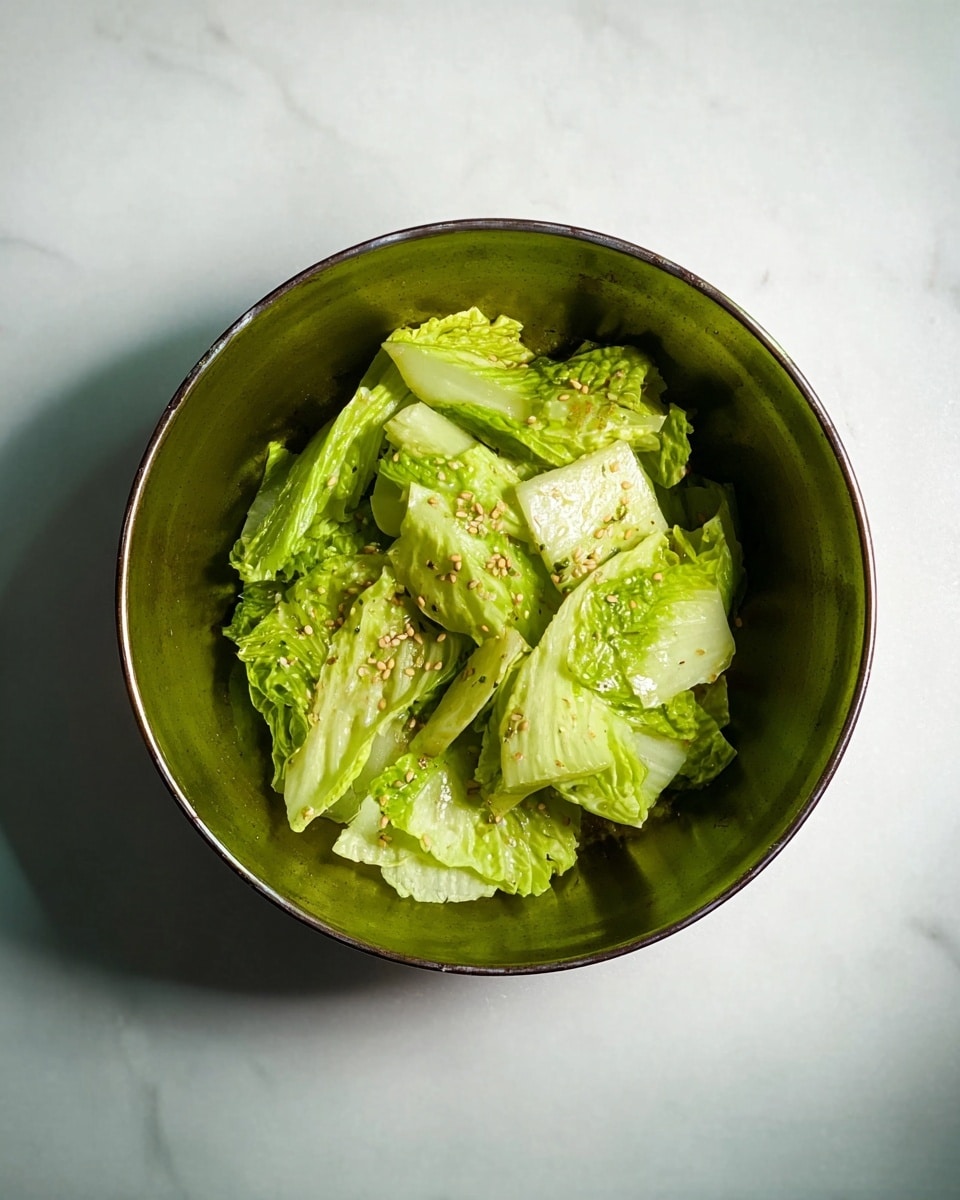 A green metal bowl holds a simple salad of light green cabbage pieces with scattered small seeds on top. The cabbage leaves have a fresh, slightly shiny texture, and they are arranged unevenly inside the bowl. The bowl sits on a white marbled surface, showing soft light and subtle shadows around it. Photo taken with an iphone --ar 4:5 --v 7