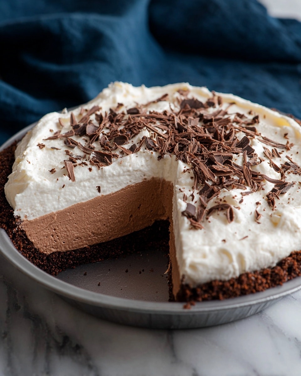 The image shows a three-layer dessert in a round metal pie tin with one large slice removed, revealing the inside. The bottom layer is a thin, dark brown crust with a crumbly texture. Above that is a thick, creamy chocolate mousse layer that is light brown and smooth. The top layer is a thick, white whipped cream, swirled on the surface with soft peaks. On top of the whipped cream are scattered dark chocolate shavings that add texture and a rich brown color contrast. The pie tin is placed on a white marbled surface, and a dark blue cloth is softly folded in the background. photo taken with an iphone --ar 4:5 --v 7