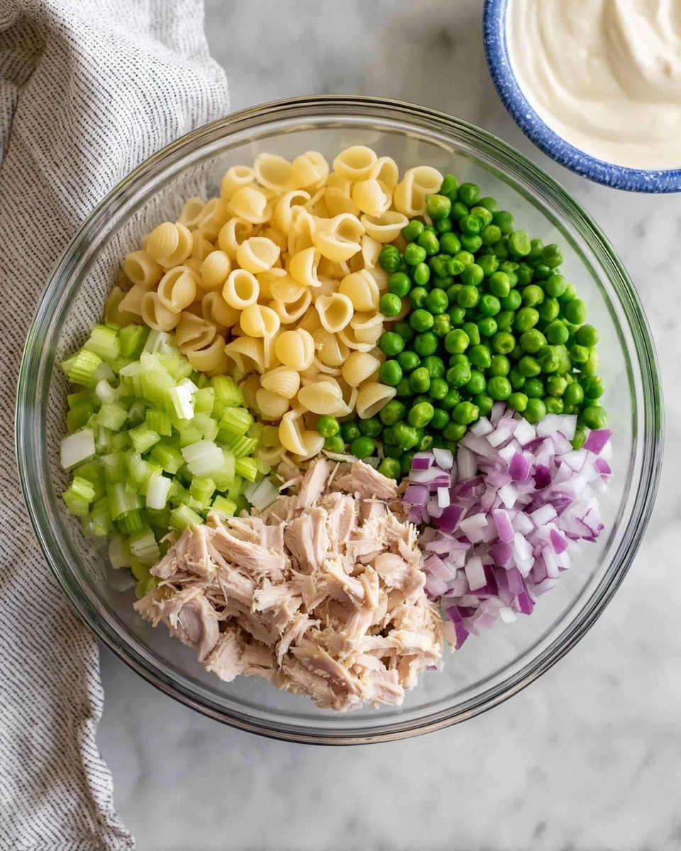 A clear glass bowl sits on a white marbled surface, filled with five distinct layers of ingredients neatly separated inside. At the top, there is a pale yellow layer of small shell pasta. To the right, bright green peas form a solid round section. Below the peas, finely chopped purple and white onion pieces create a small mound. On the left below the pasta, diced light green celery occupies a quarter of the bowl. At the bottom left, light brown shredded cooked chicken fills the remaining section. Next to this bowl, a white bowl with a blue rim holds a smooth off-white creamy sauce. A light gray and white striped cloth is partially visible in the upper left corner of the frame photo taken with an iphone --ar 4:5 --v 7