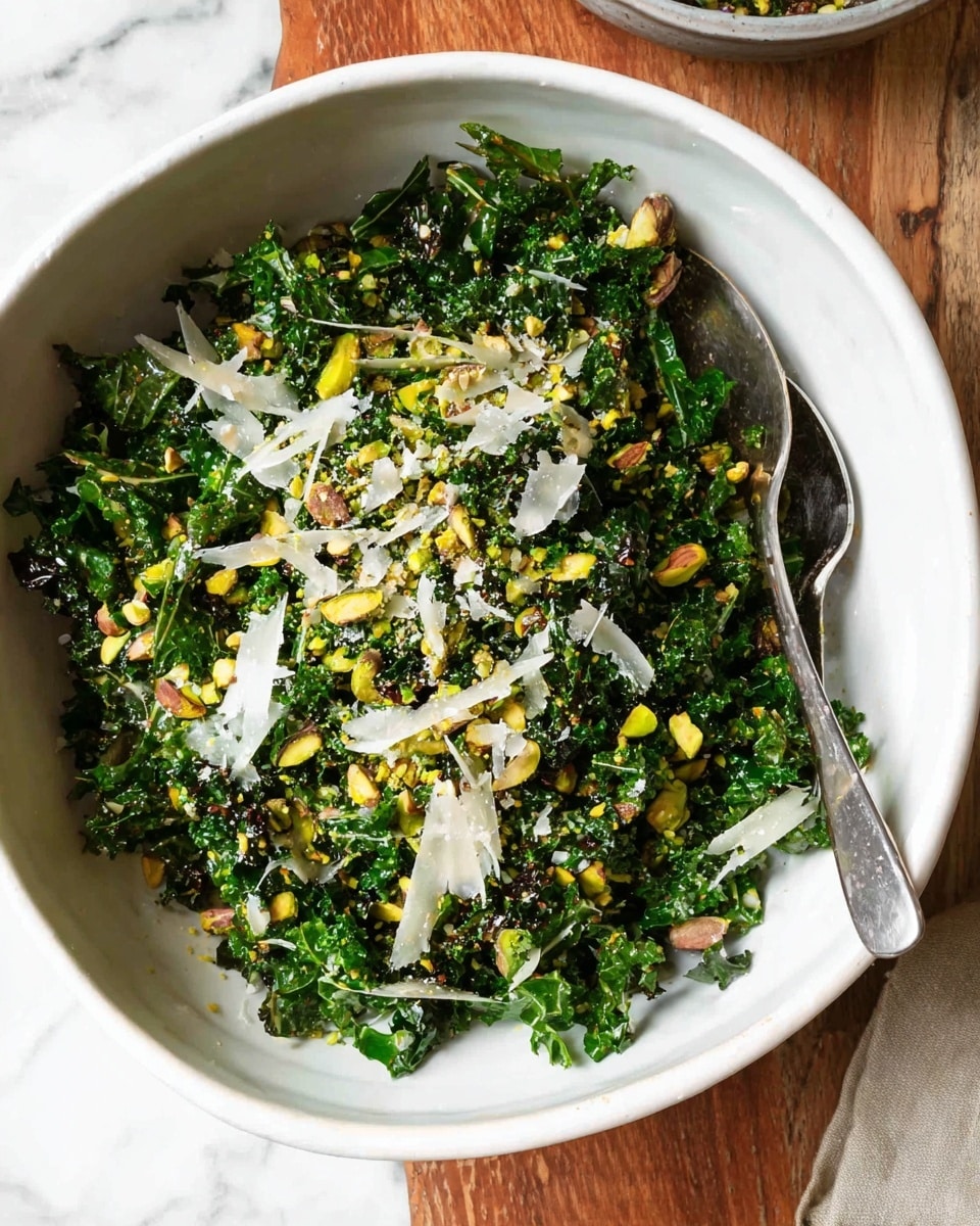 A white bowl filled with a green kale salad covered with small chopped pistachios, and thin white shavings of cheese sprinkled all over. The kale leaves look fresh and slightly glossy, mixed with bits of crushed pistachio nuts that add rough texture and greenish-brown colors. There is a silver spoon partially inside the bowl on the right side. The bowl is placed on a wooden surface with a white marbled background visible behind it. Photo taken with an iphone --ar 4:5 --v 7