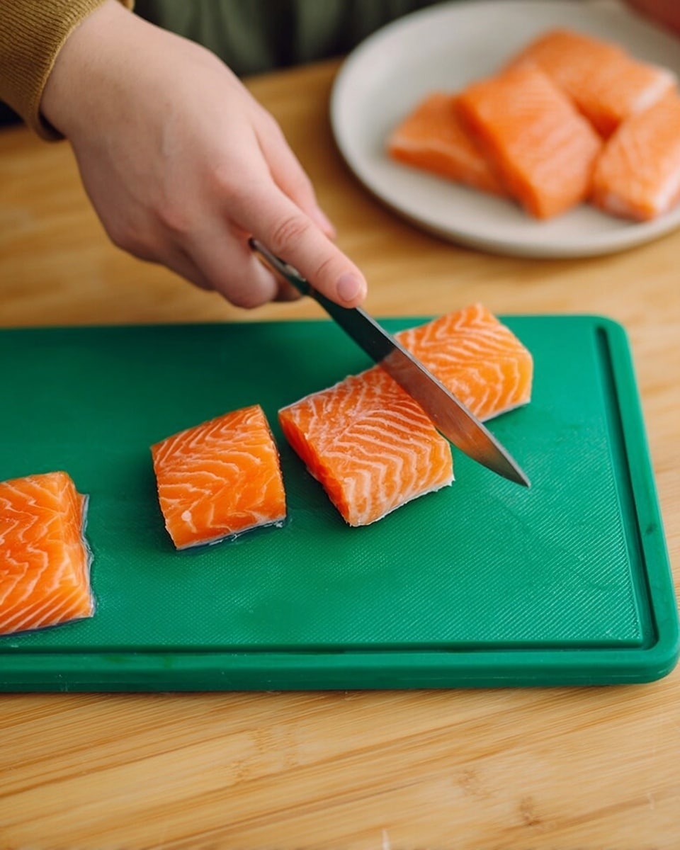 A close-up view of a woman's hand holding a knife slicing a fresh orange piece of salmon into three smaller rectangular pieces on a green chopping board placed on a light wood surface. Another chunk of whole salmon lies at the bottom of the board, and on the top right corner, a white plate holds several more uncooked salmon pieces with orange and white marbled texture. The scene looks like a kitchen preparation moment. photo taken with an iphone --ar 4:5 --v 7