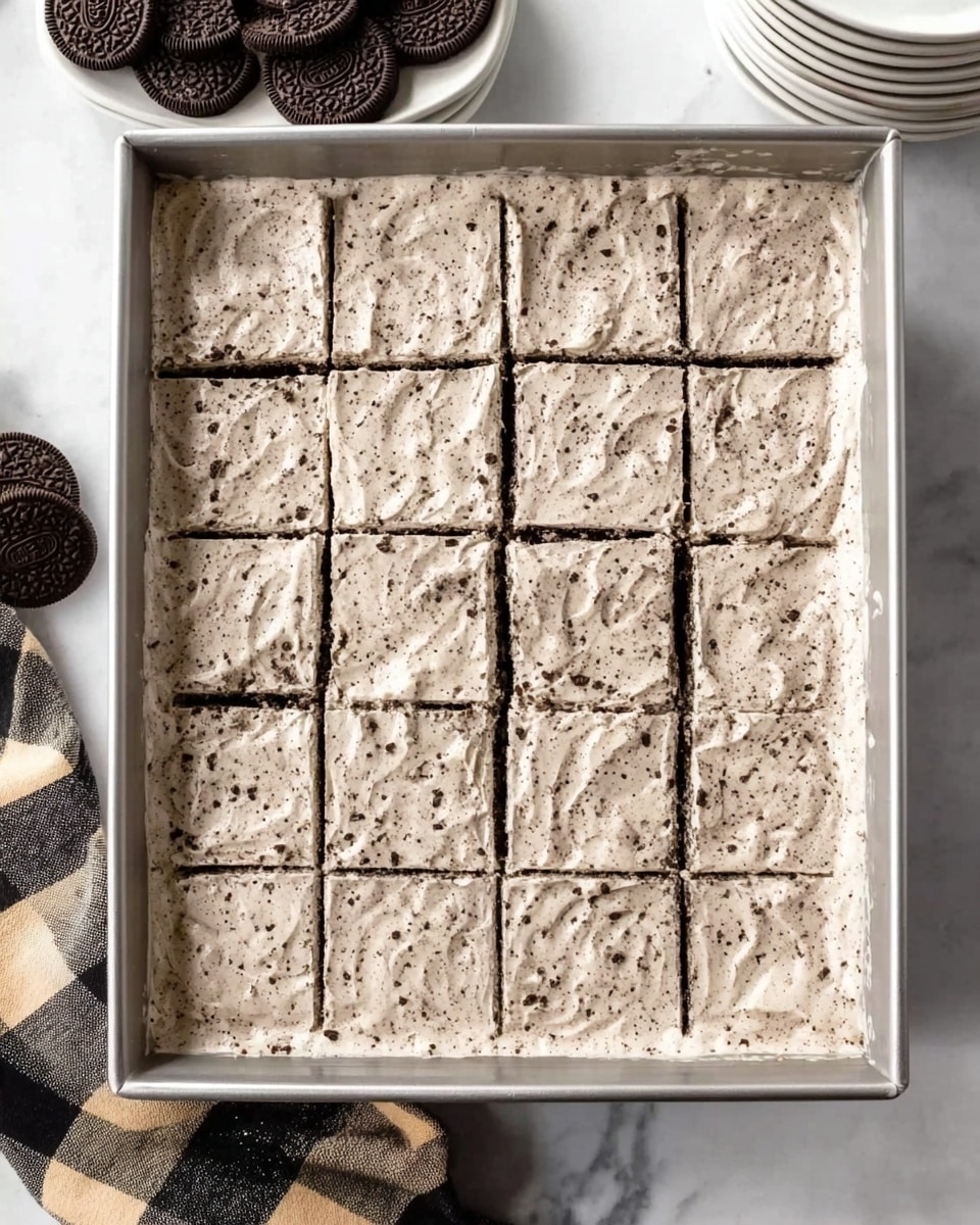 The image shows a rectangular metal baking pan filled with a thick, creamy dessert layer that has small dark specks evenly spread throughout, creating a cookies-and-cream look. The dessert is roughly divided into sixteen even squares, with clean, straight lines marking each piece. The top layer is smooth but has some gentle swirls, giving it a soft, fluffy texture. The pan is placed on a white marbled surface, with a black and beige checkered cloth partially visible on the left side. There are stacks of white plates in the background and some chocolate sandwich cookies placed near the top left corner. Photo taken with an iphone --ar 4:5 --v 7