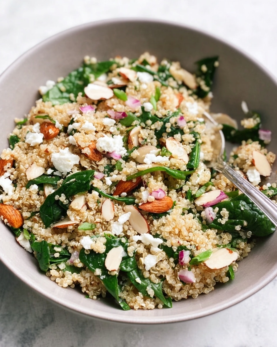 A close-up image of a light gray bowl filled with a quinoa salad. The salad has small, grainy light beige quinoa as the base layer, mixed with thinly sliced dark green spinach scattered evenly on top. Bright pink small onion cubes and golden roasted almond slices are mixed throughout. There are small white crumbles of cheese sprinkled around the salad. A metal fork is placed inside the bowl, touching the salad on the right side. The bowl sits on a white marbled surface. photo taken with an iphone --ar 4:5 --v 7
